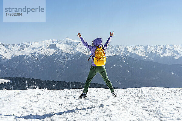 Junge glückliche Frau mit gelbem Rucksack springt in verschneite Berge