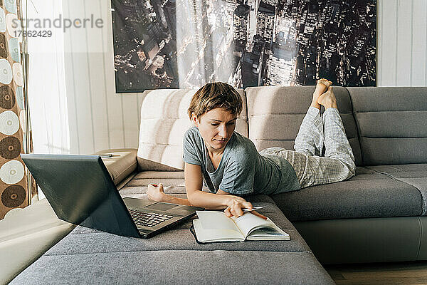 Eine Frau schaut sich Notizen in einem Notizbuch an und benutzt dazu einen Laptop  der auf der Couch liegt