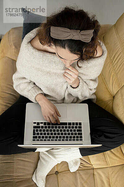 Draufsicht auf eine junge Frau  die mit gekreuzten Beinen auf dem Sofa sitzt und am Laptop arbeitet