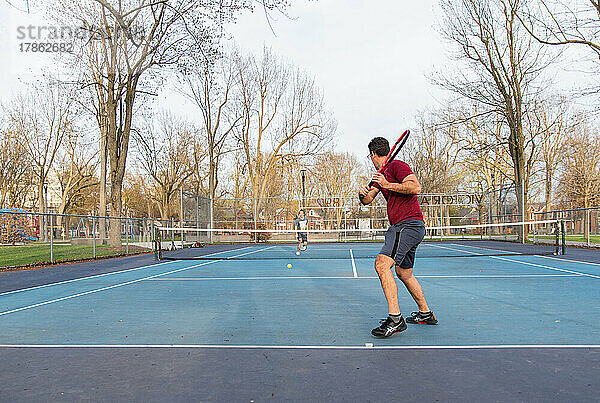 Mann spielt im Frühling mit seinem jugendlichen Sohn Tennis auf einem Hartplatz im Freien.