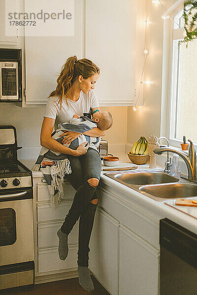 Mutter hält Säugling  während sie in kleiner Küche auf der Theke sitzt