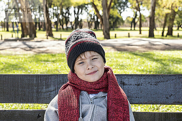 Kind sitzt auf einer Bank im Park und trägt eine Wollmütze und einen Schal