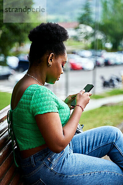 Afroamerikanische Frau sitzt mit Handy auf Bank im Park