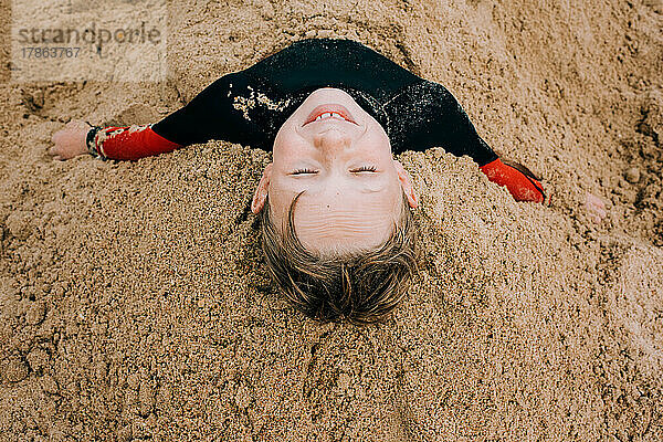 Junge liegt glücklich im Sand vergraben und hat Spaß am Strand