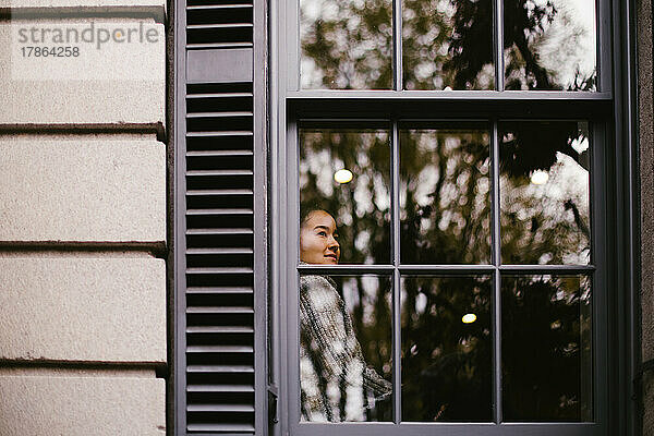 Asiatische Frau blickt zufrieden aus dem komfortablen Haus aus dem Fenster