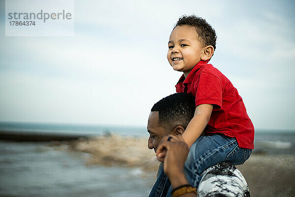 Mischlingssohn lächelt glücklich auf den Schultern des schwarzen Vaters am Strand