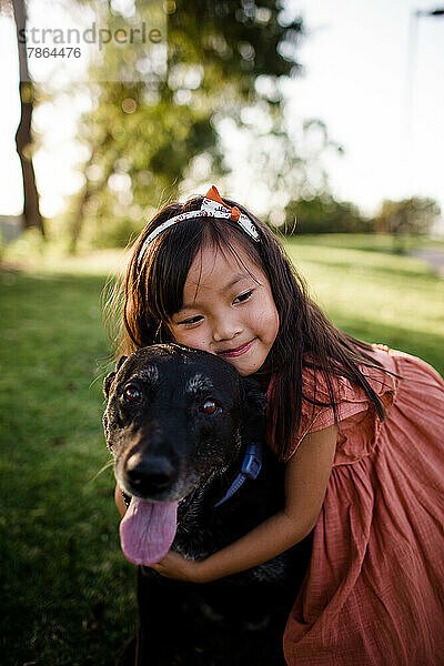 Sechs Jahre altes asiatisches Mädchen umarmt schwarzen Labrador im Park in San Diego