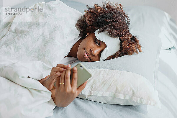 Afrikanerin mit Afrohaar im Bett mit Smartphone. Schlafmaske