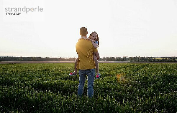 Vater trägt seine Tochter  während er bei Sonnenuntergang auf einem Feld spaziert