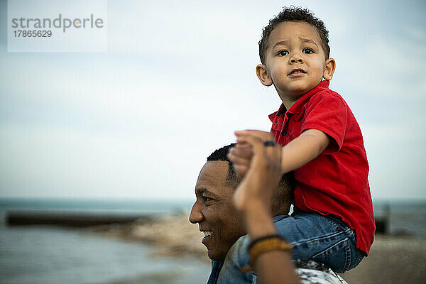Schwarzer Vater lässt dreijährigen Sohn auf Schultern am Wasser reiten