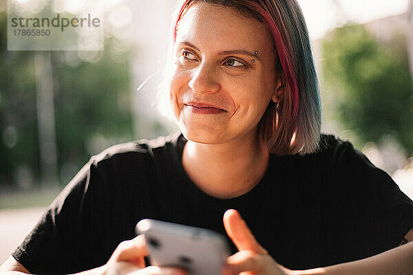 Glückliche junge Frau lächelt  schaut weg und hält ihr Smartphone im Freien