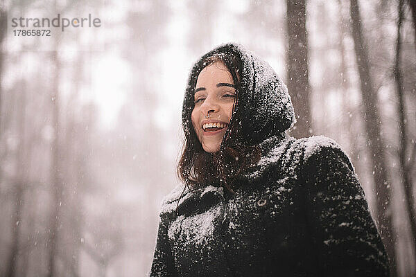 Porträt einer glücklichen jungen Frau  die bei Schneefall im Wald steht