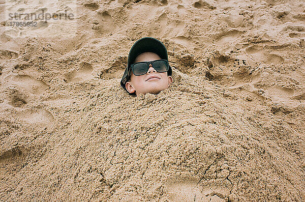 Im Sand begrabener Junge entspannt sich glücklich am Strand