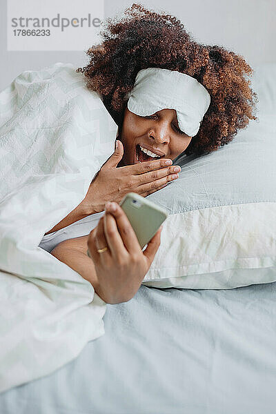 Afrikanerin gähnt im Bett und schaut sich Nachrichten am Telefon an