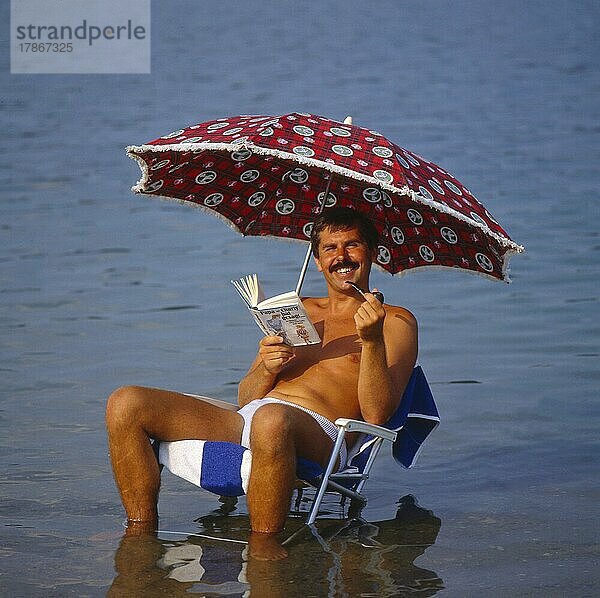 Junger Mann mit Sonnenstuhl und Sonnenschirm am Strand  mit Pfeife rauchend  im Meer  Urlaub  Erholung  young man with solar chair and sunshade on the beach  with whistle smoking  in the sea  vacation  rest