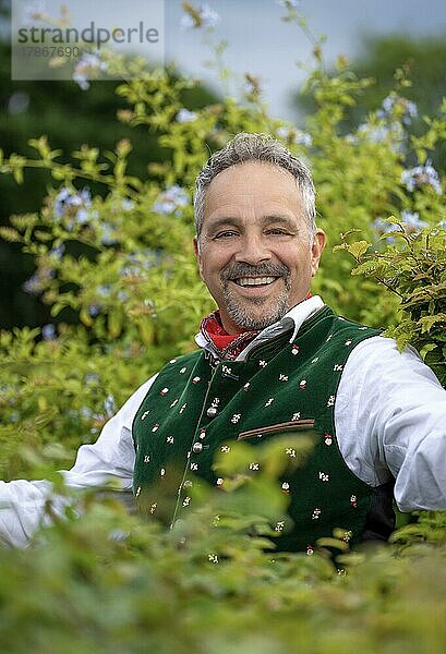 Lachender Mann im mittleren Alter in Bayrischer Tracht steht an Hecke  Karlsruhe  Deutschland  Europa