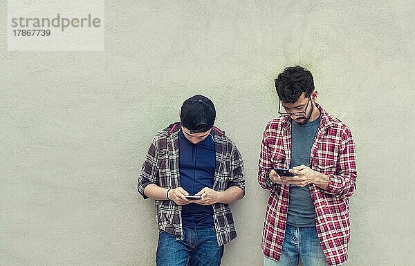 Zwei Teenager-Freunde an einer Wand  die ihre Handys überprüfen  Zwei Freunde  die an einer Wand lehnen und auf ihren Handys simsen. Freund zeigt seinem Freund sein Handy  Lächelnde Freunde prüfen ihre Handys