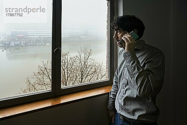 Latino-Geschäftsmann  der in einem Büro mit Blick auf den Fluss arbeitet. Er telefoniert  während er aus dem Fenster schaut