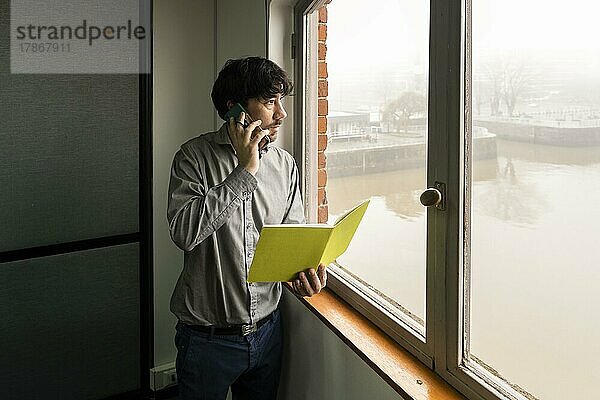 Latino-Geschäftsmann bei der Arbeit in einem Büro mit Blick auf den Fluss. Er telefoniert und prüft Notizen  während er aus dem Fenster schaut