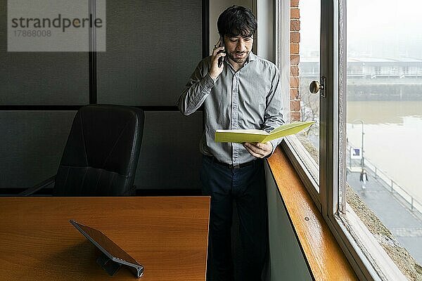 Latino-Geschäftsmann bei der Arbeit in einem Büro mit Blick auf den Fluss. Er telefoniert und geht Notizen durch