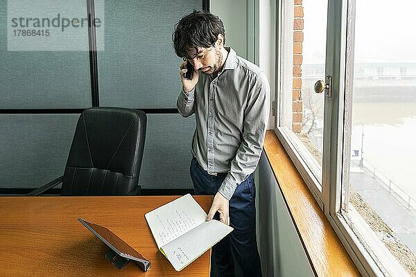 Latino-Geschäftsmann bei der Arbeit in einem Büro mit Blick auf den Fluss. Er telefoniert und geht Notizen durch
