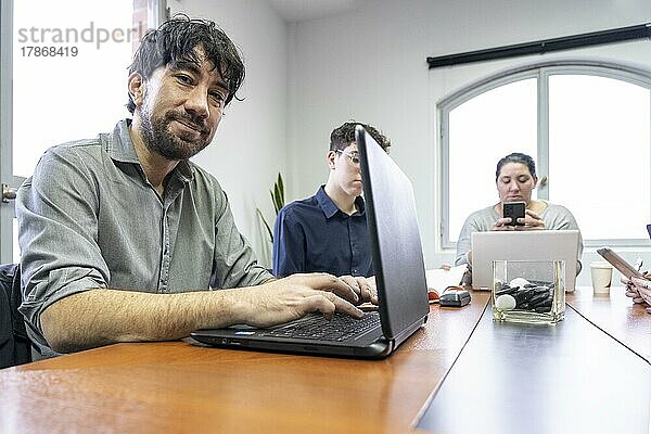 Eine Gruppe von Angestellten sitzt um den Besprechungstisch im Büro und arbeitet an ihren Notebooks  einer von ihnen schaut in die Kamera