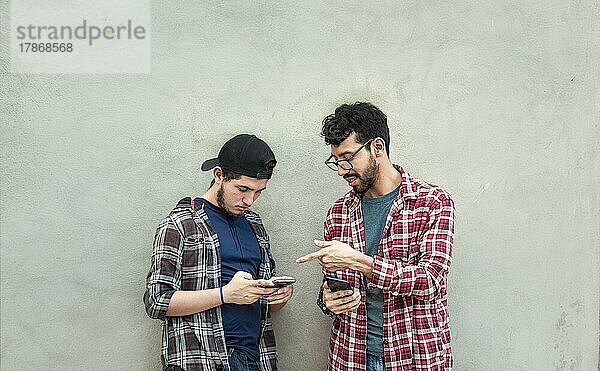 Zwei junge Freunde  die an einer Wand lehnen und ihre Handys überprüfen  Zwei Freunde  die an einer Wand lehnen und auf den Inhalt ihrer Handys schauen. Freund zeigt seinem Freund im Freien sein Handy