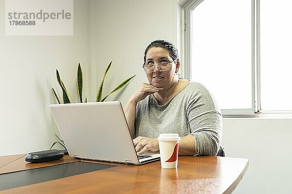 Reife Geschäftsfrau sitzt am Kopfende eines Tisches in einem Besprechungsraum  arbeitet an ihrem Computer und sieht jemanden an  der außerhalb des Bildes steht