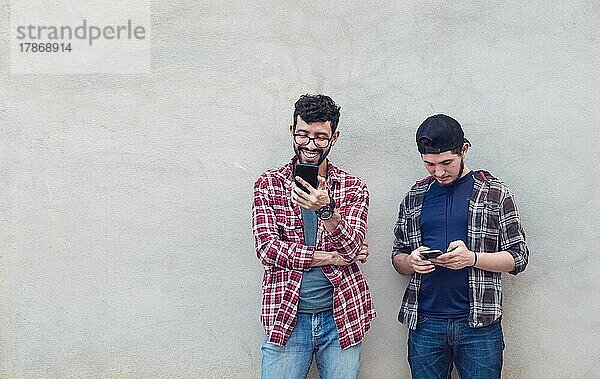 Zwei lächelnde Freunde  die an einer Wand lehnen und ihre Handys überprüfen  Freunde  die an einer Wand lehnen und auf ihren Handys simsen. Freund zeigt seinem Freund sein Handy  Lächelnde Freunde prüfen Handys