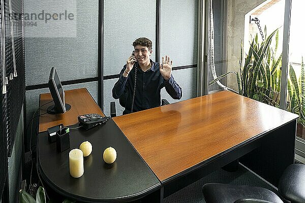 Geschäftsmann sitzt an seinem Schreibtisch und telefoniert  schaut in die Kamera und winkt