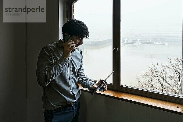 Latino-Geschäftsmann bei der Arbeit in einem Büro mit Blick auf den Fluss. Er telefoniert und geht Notizen durch