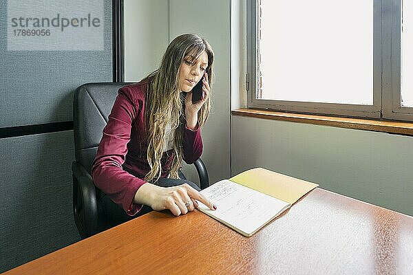 Junge blonde Geschäftsfrau sitzt in ihrem Büro und telefoniert  während sie Notizen überprüft