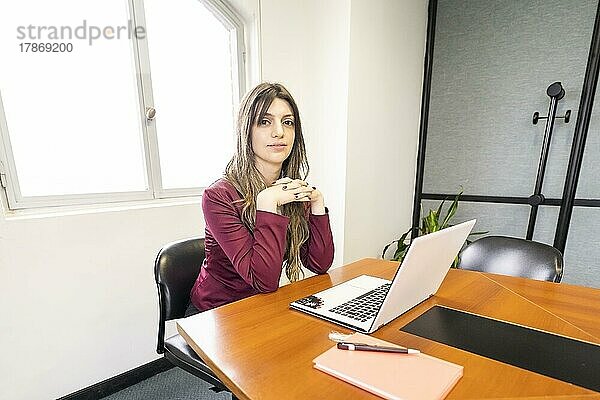 Junge blonde Geschäftsfrau  die am Kopfende des Tisches im Bürobesprechungsraum sitzt  mit ihrem Computer und Handy  und in die Kamera schaut