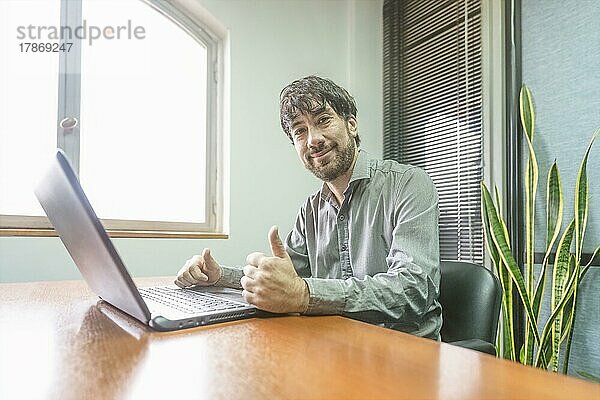 Geschäftsmann  der an seinem Schreibtisch sitzt und an seinem Notebook arbeitet  lächelnd und mit erhobenem Daumen in die Kamera schaut