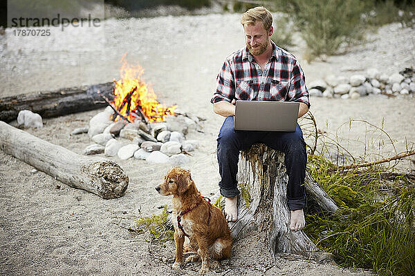 Lächelnder Geschäftsmann sitzt mit Hund am Lagerfeuer und benutzt Laptop