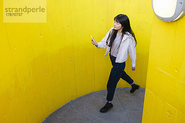 Junge Frau benutzt Mobiltelefon und läuft zwischen gelben Wänden entlang