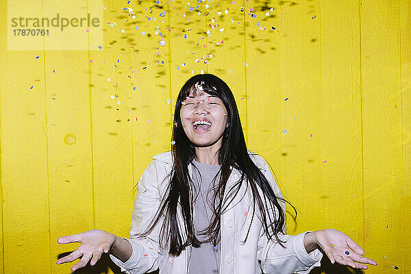 Fröhliche junge Frau steht unter Konfetti vor gelber Wand