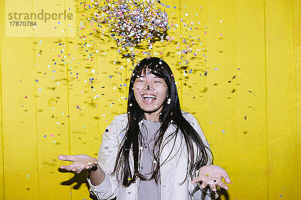 Fröhliche junge Frau wirft Konfetti vor gelbe Wand