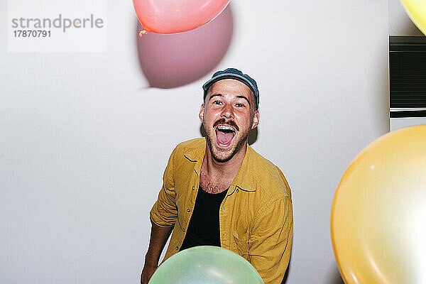 Fröhlicher Mann genießt es mit bunten Luftballons vor der Wand zu Hause