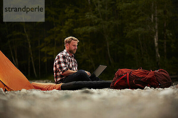 Businessman using laptop sitting on inflatable mattress at campsite