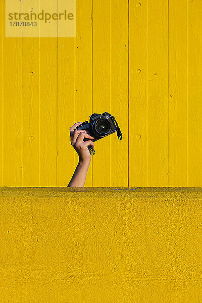Hand einer Frau hält Kamera hinter gelber Wand