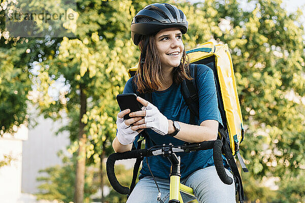 Lächelnde Lieferfrau mit Helm sitzt mit Smartphone auf dem Fahrrad