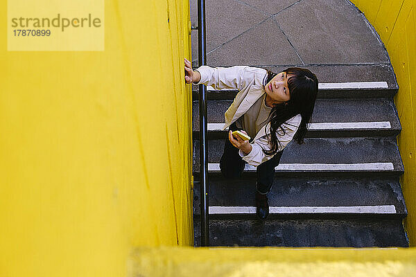 Junge Frau mit Smartphone auf der Treppe und schaut nach oben