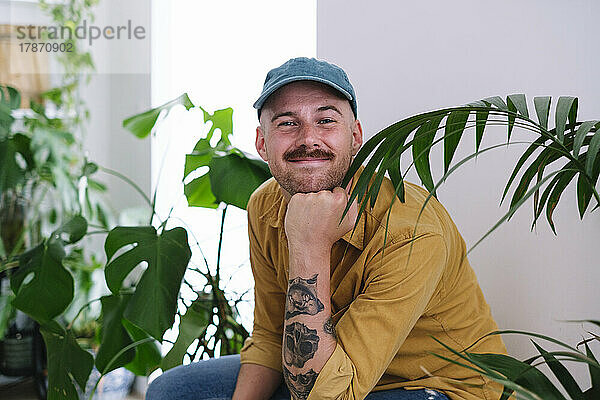 Happy man with hand on chin sitting amidst plants at home