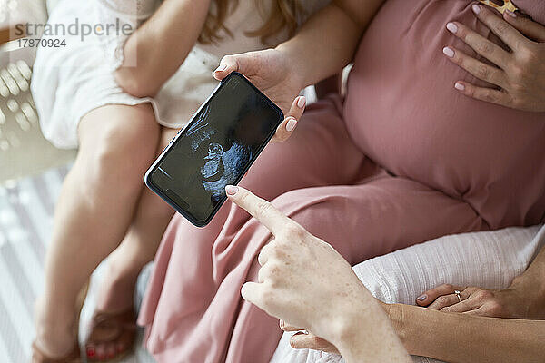 Hand einer schwangeren Frau  die Freunden das Ultraschallergebnis auf dem Smartphone zeigt