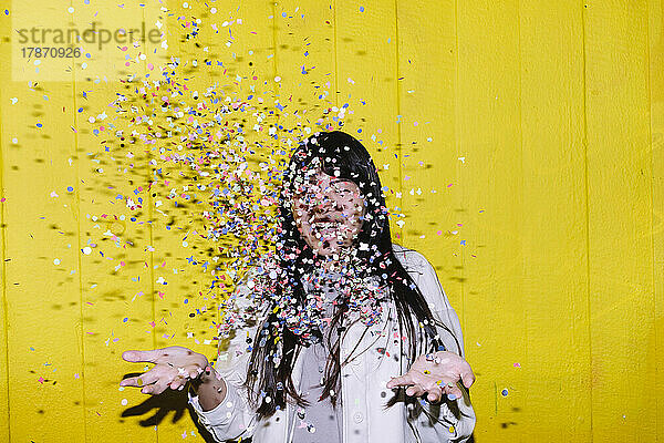 Glückliche Frau wirft buntes Konfetti vor gelbe Wand