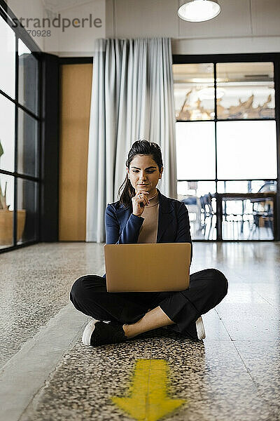 Junge Geschäftsfrau sitzt mit der Hand am Kinn und schaut im Büro auf den Laptop