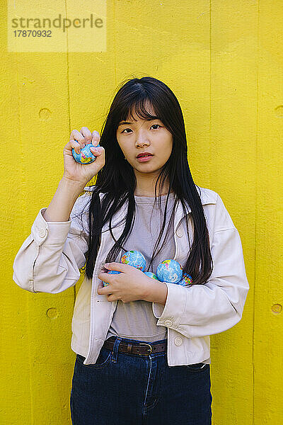 Young woman holding globe standing in front of yellow wall