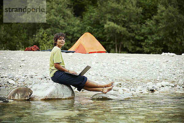 Lächelnde Geschäftsfrau mit Laptop sitzt auf einem Felsen am Flussufer