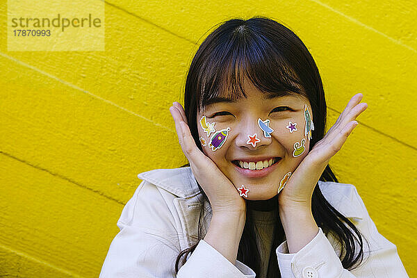 Happy young woman with stickers on face in front of yellow wall